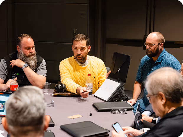 Group of men having a discussion around a table, one man in a yellow sweater showing a document in a black folder.