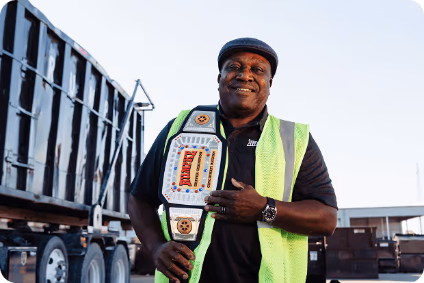 Smiling man wearing a neon yellow safety vest and a black cap holding a championship belt outdoors near trucks.