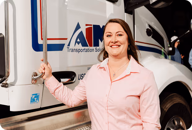 A smiling woman in a light pink shirt stands beside a white truck with AIM Transportation Solutions logo on the door.