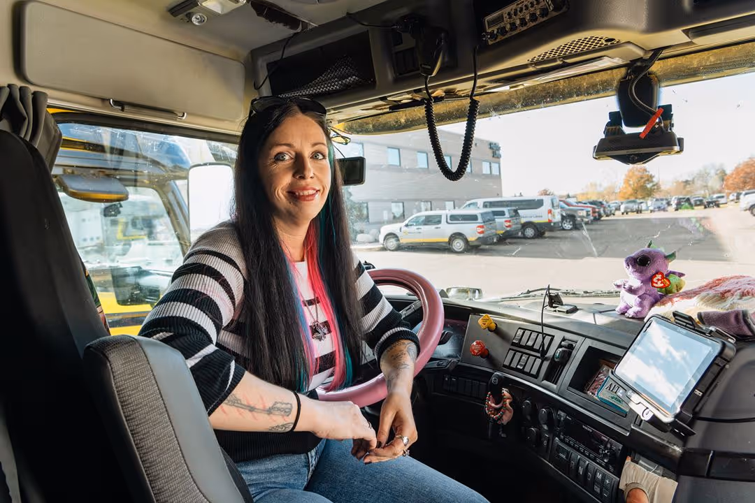 Woman with long dark hair and tattoos sitting in the driver's seat of a large vehicle, smiling at the camera.
