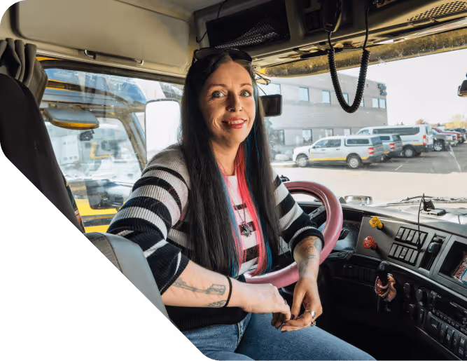 Woman with long dark hair and tattoos sitting in the driver's seat of a large vehicle, smiling at the camera.