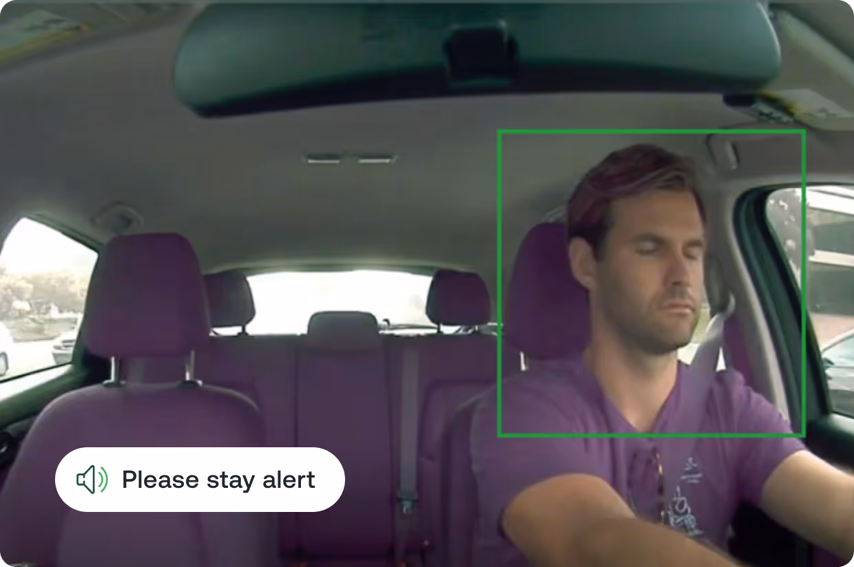 Driver with eyes closed behind the wheel inside a car with a safety message saying 'Please stay alert'.