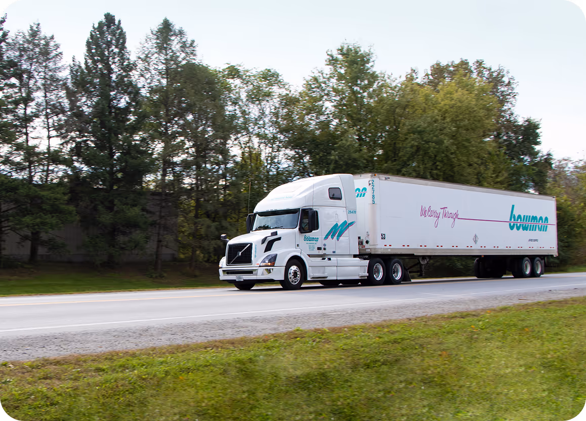 White semi-truck with Boomer logo and slogan 'We Carry Through' driving on a road with trees in the background.