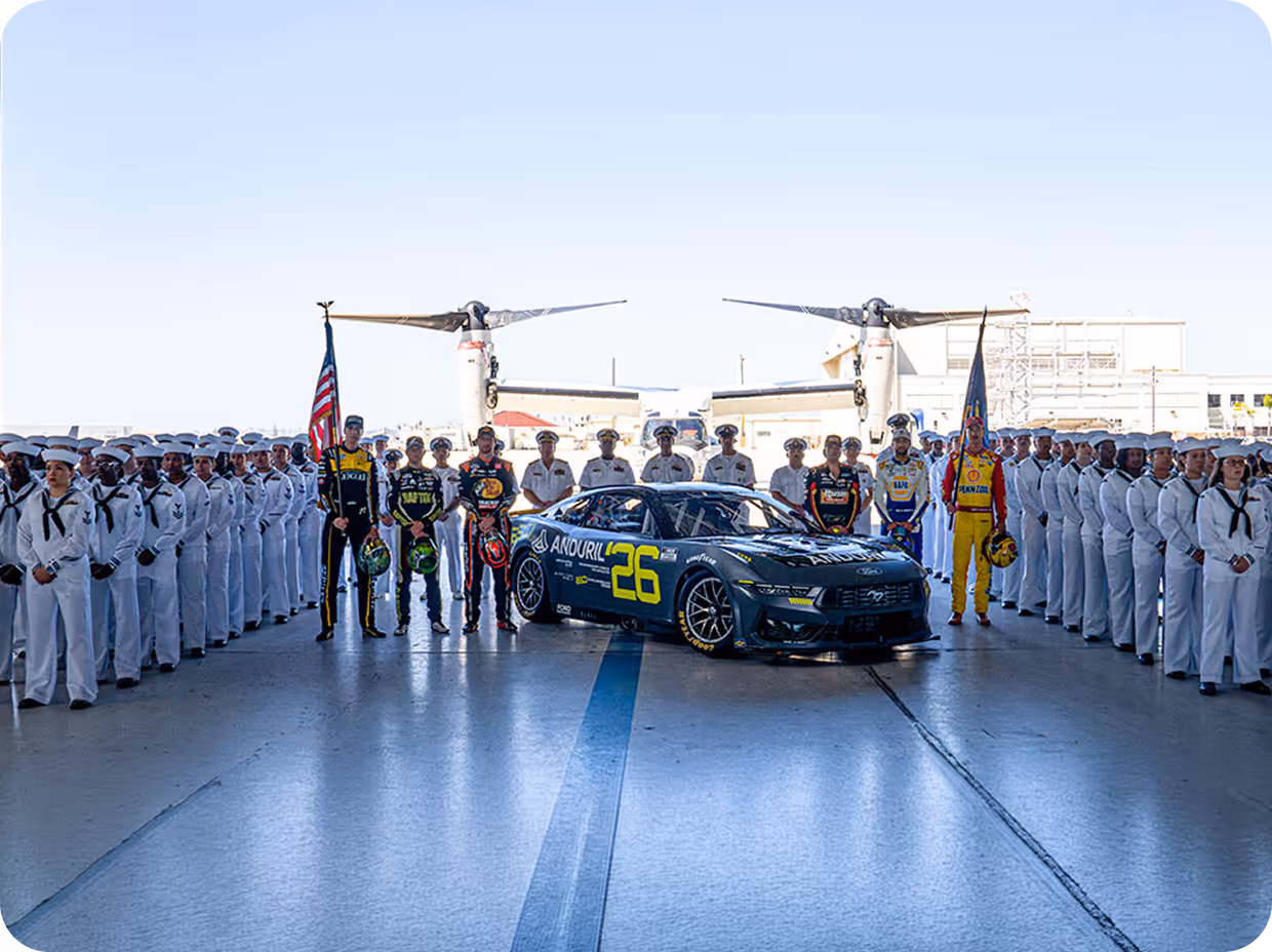 Race car number 26 surrounded by uniformed navy personnel and race drivers holding flags in a hangar with aircraft visible in the background.