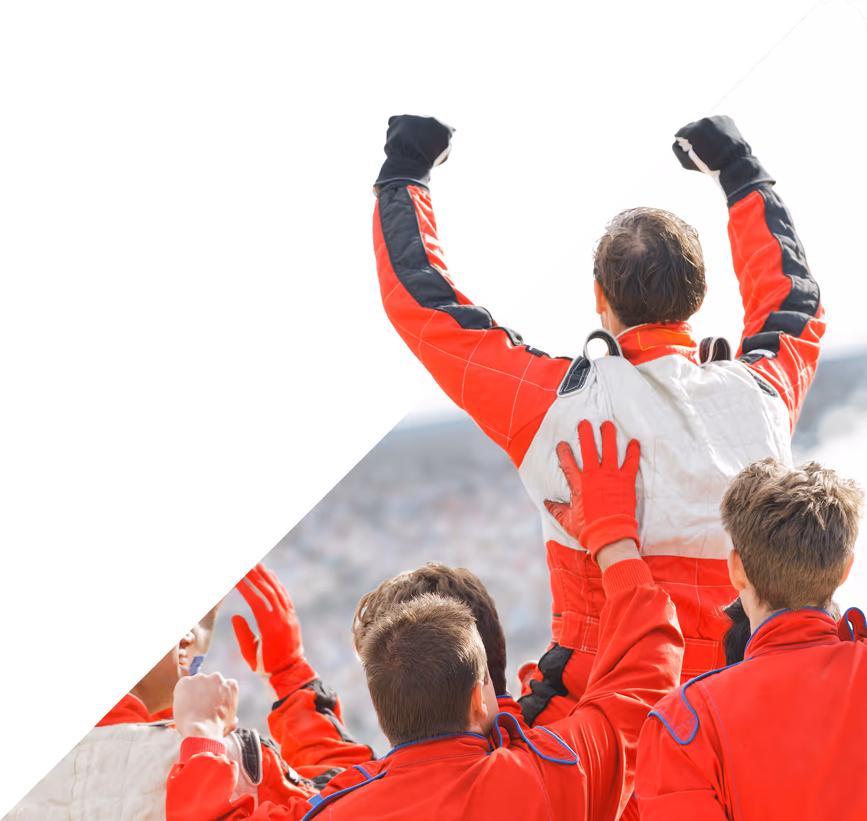 A race car driver in red and white gear is lifted by his cheering team, raising his fists in victory.