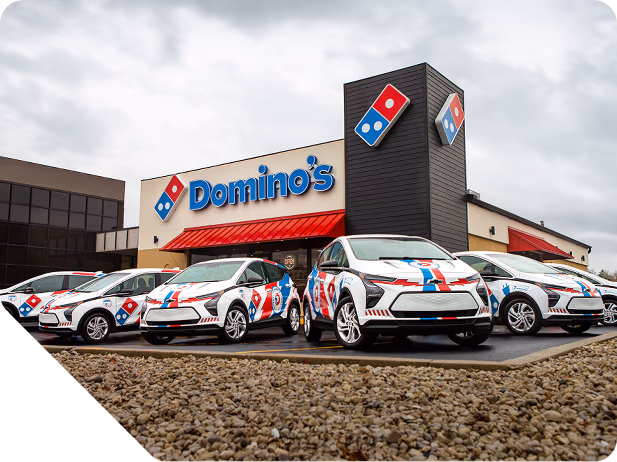 Domino's pizza store front with several white delivery cars featuring Domino's logo parked outside under an overcast sky.