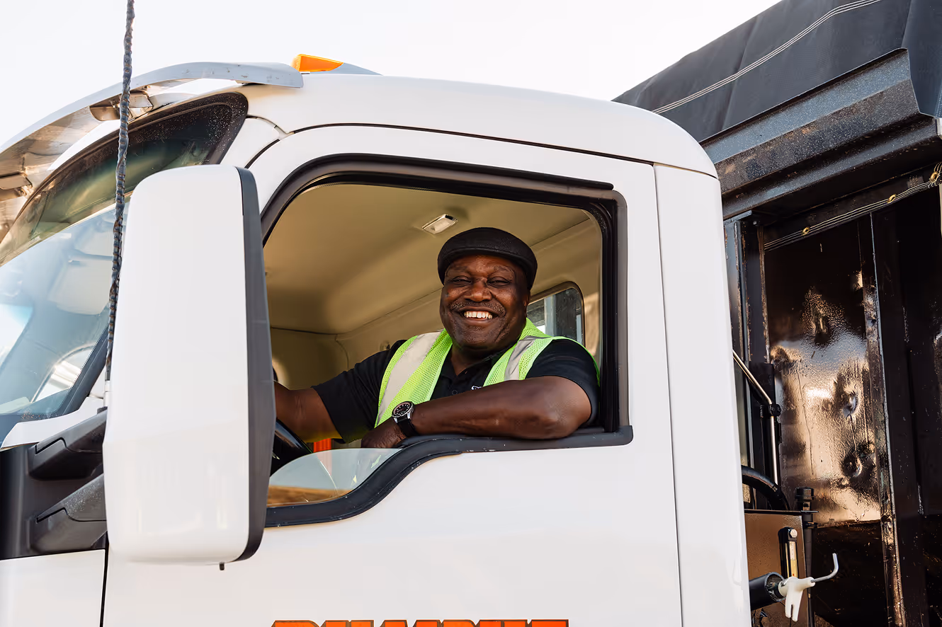 Smiling truck driver wearing a green safety vest sitting in the driver's seat of a white truck.