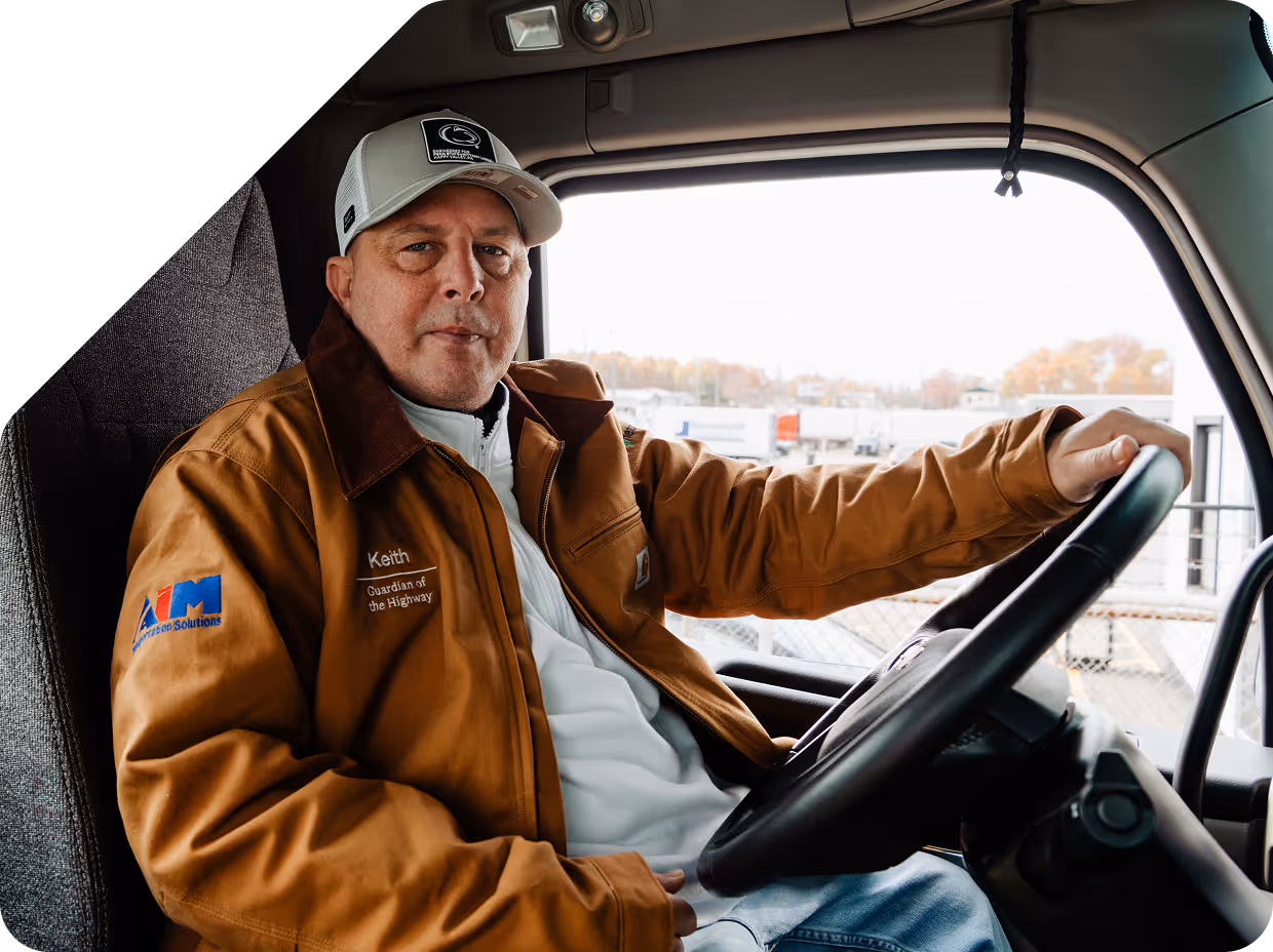 Man wearing a brown jacket and white cap sitting in the driver's seat of a truck, holding the steering wheel.