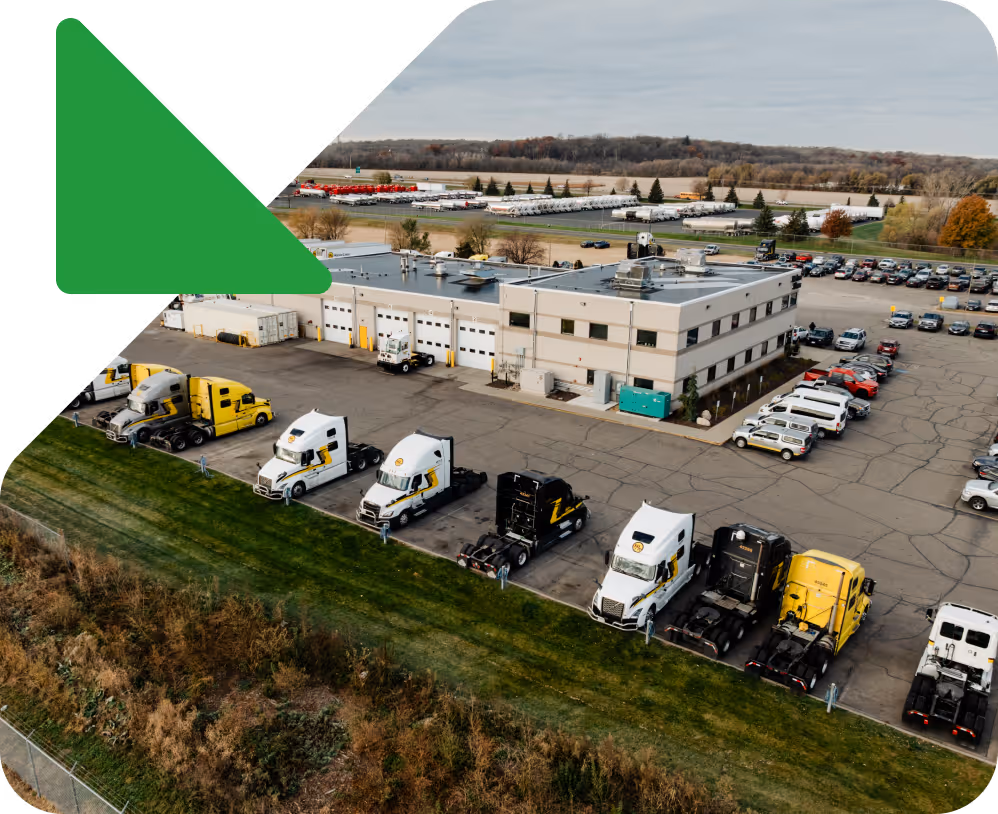 Aerial view of a commercial warehouse with trucks and vehicles parked around it in a paved lot with open fields and trees in the background.