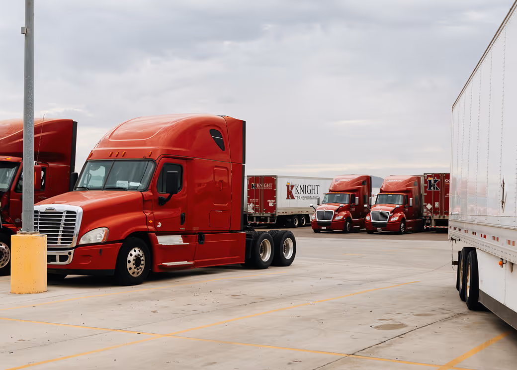 Red Knight Transportation semi trucks parked on concrete lot under cloudy sky.