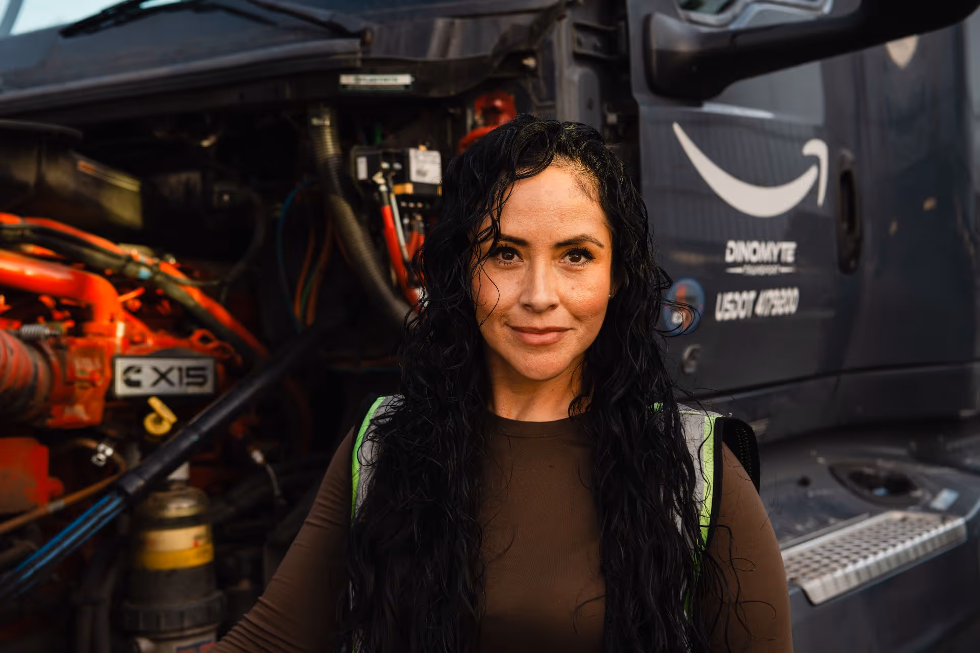 Woman with long dark hair wearing safety vest stands in front of a truck with open hood.