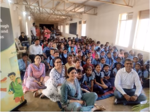 Group of children and adults sitting on the floor in a large room with windows and a banner.