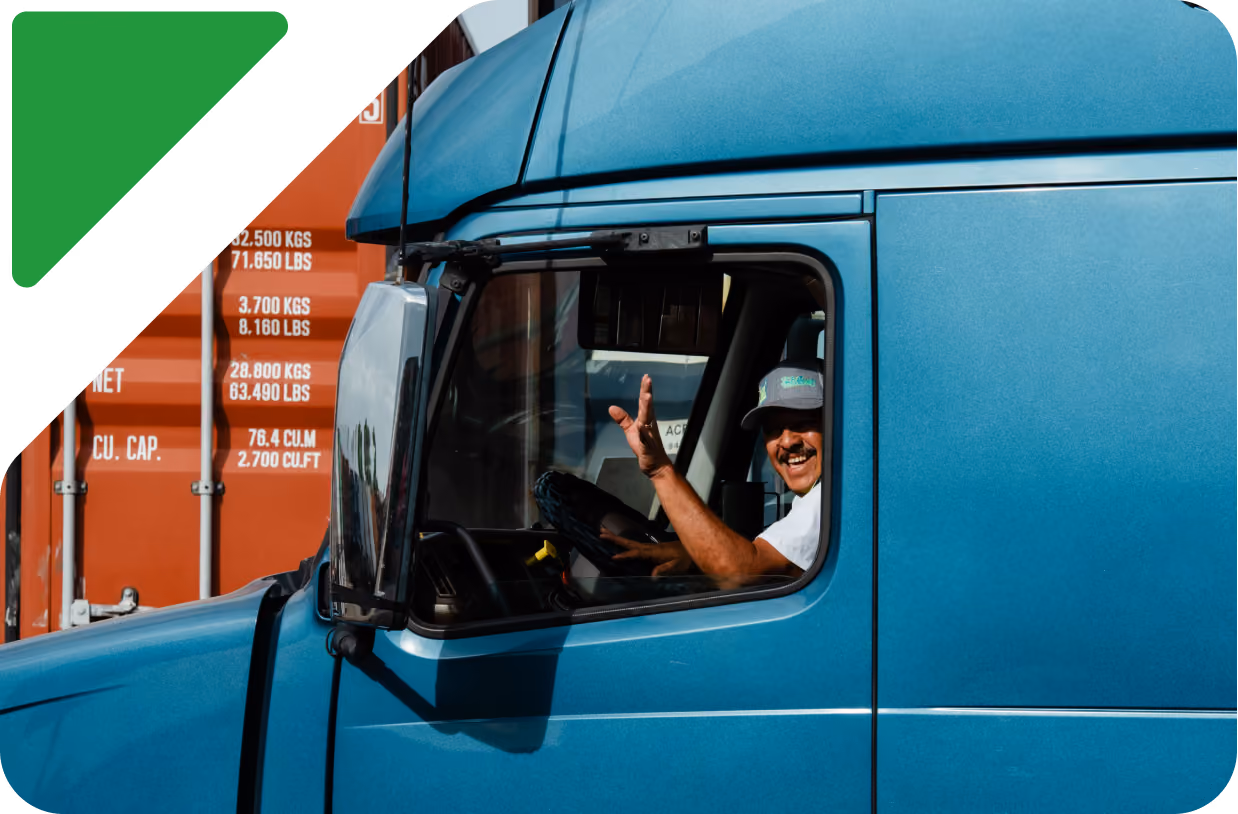 Smiling man wearing a cap waves from the driver's seat of a blue semi-truck cab in front of orange shipping containers.