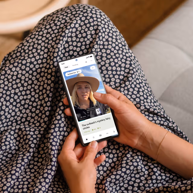 Person holding a smartphone displaying a video of a blonde woman wearing a wide-brimmed hat with the headline 'The outback's mystery fairy circles'.