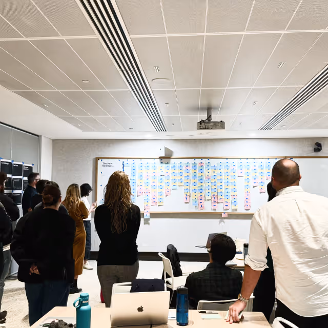 Group of people standing and sitting in a meeting room looking at a large whiteboard covered with organized colorful sticky notes.