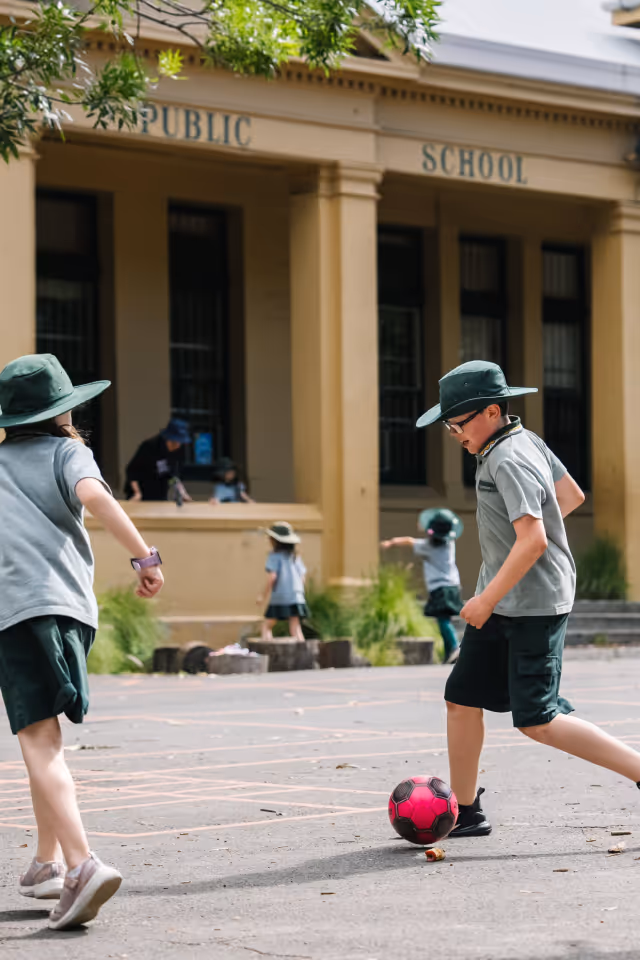 Children wearing hats and school uniforms playing with a pink soccer ball on a school playground.