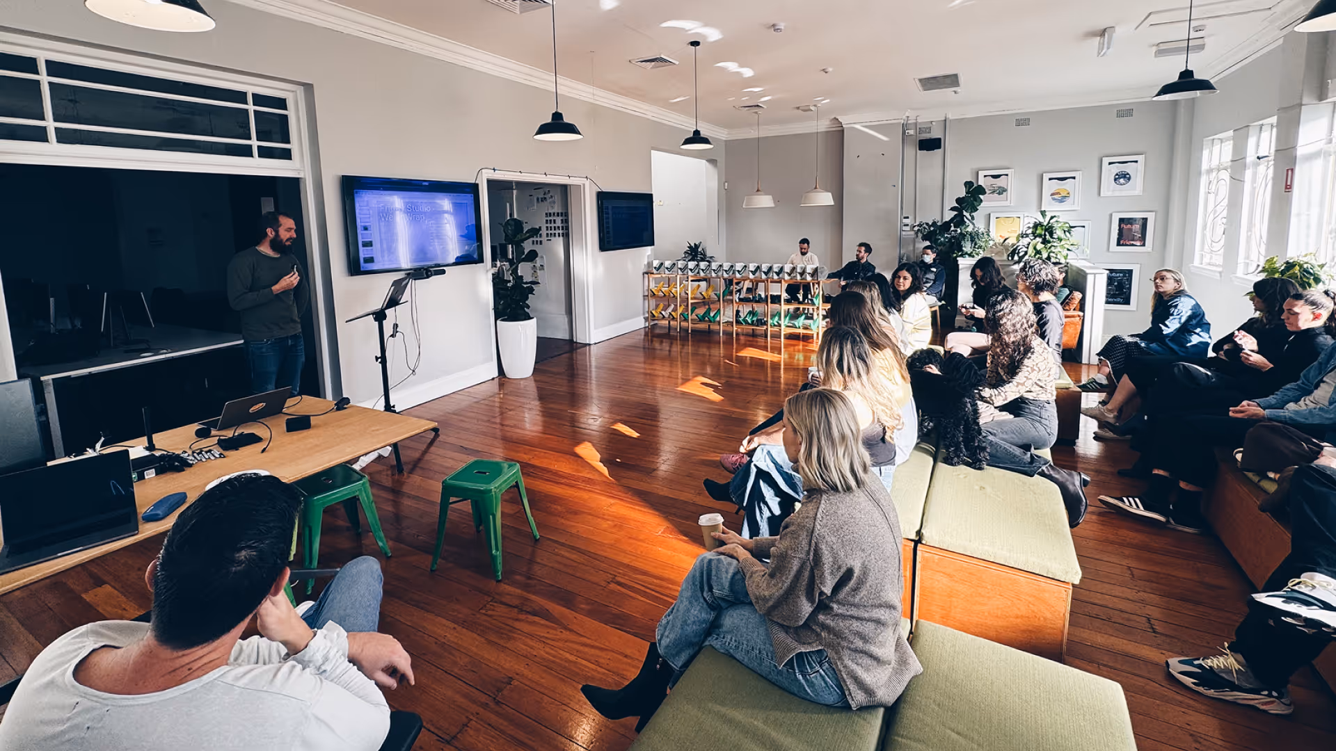 Man giving a presentation to a seated audience in a bright room with wooden floors and green cushioned benches.