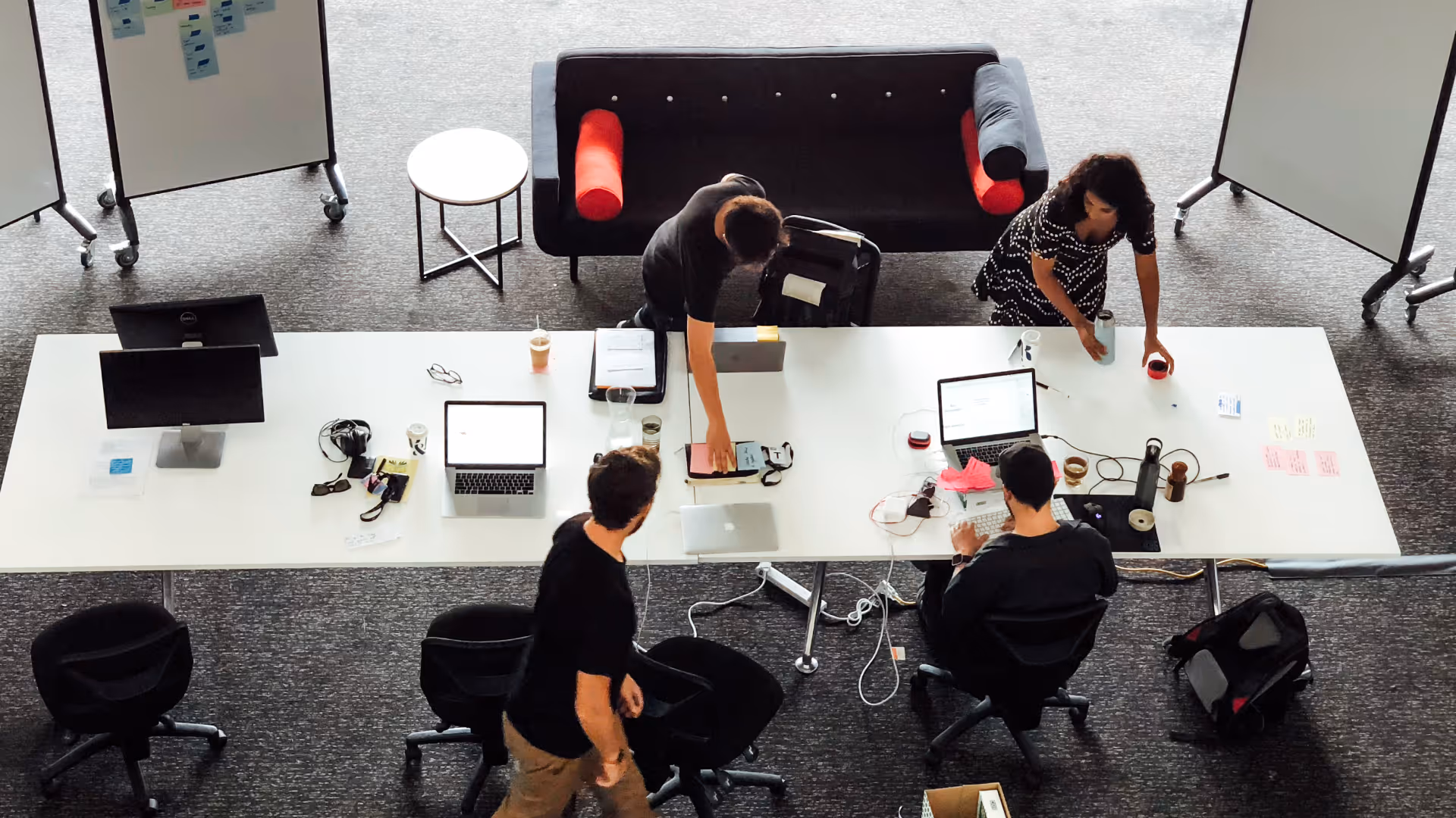 Overhead view of four people working at a large white office table with laptops, monitors, and office supplies, with two whiteboards and a black sofa in the background.