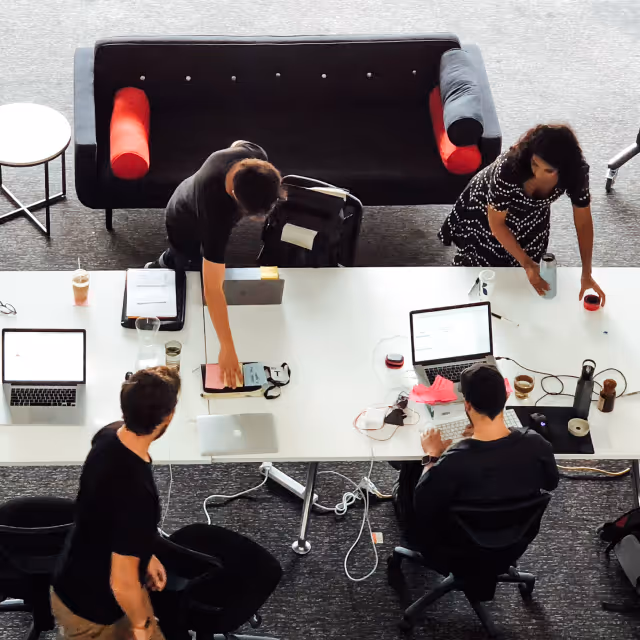 Top-down view of four people working at a white table with laptops, drinks, and office supplies in a modern workspace near a black sofa with red cushions.