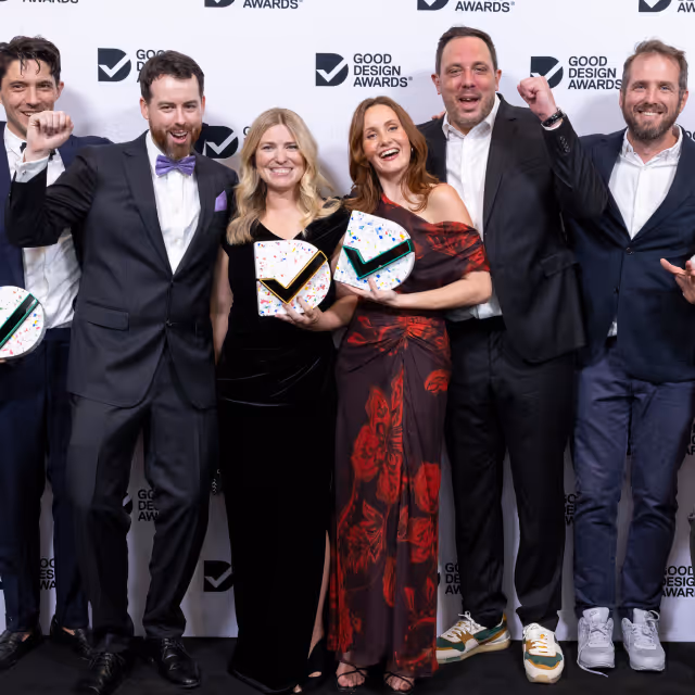 Six people dressed formally posing and smiling at Good Design Awards event, with two women holding award trophies.