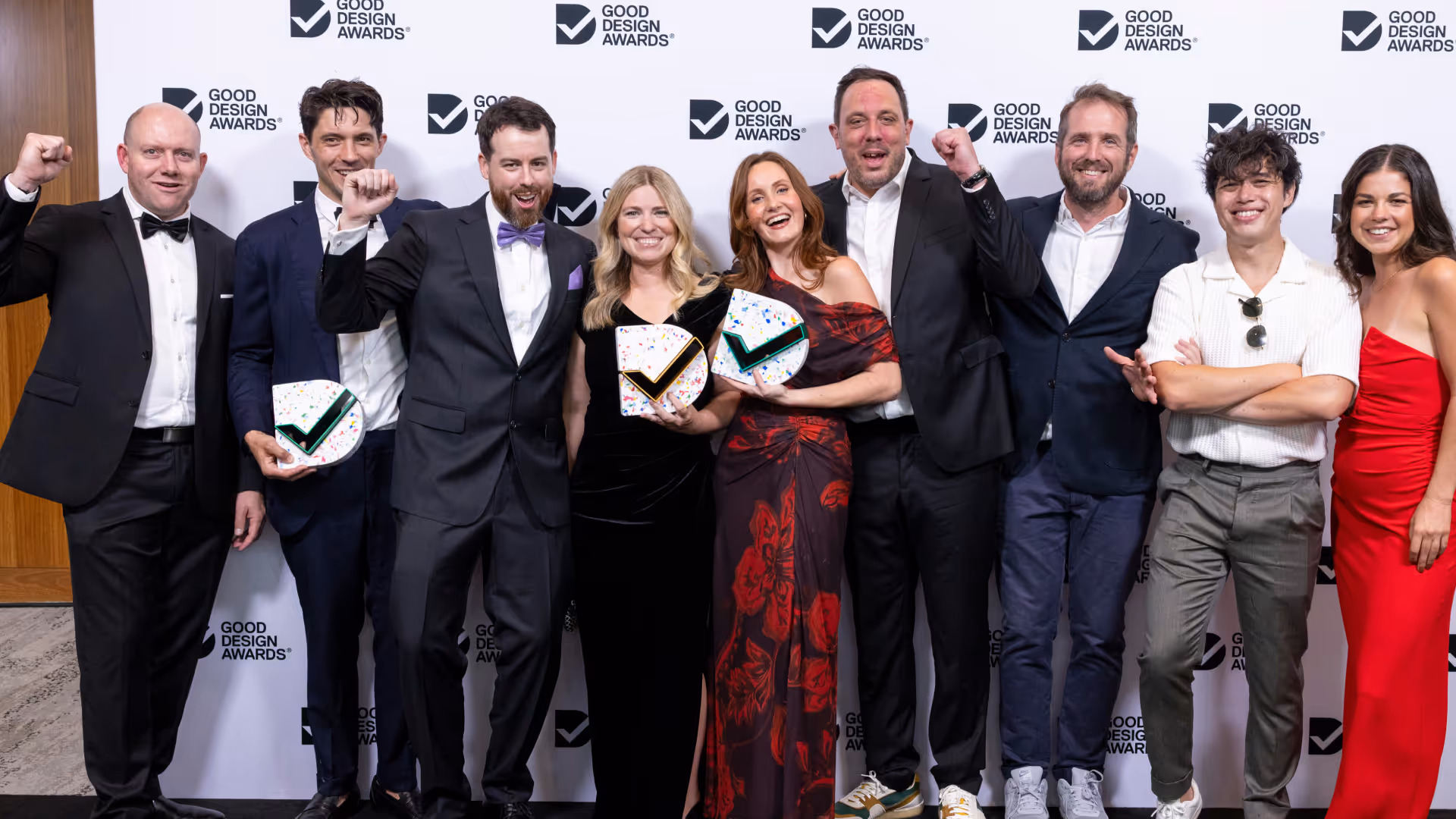Eight people dressed in formal and semi-formal attire celebrating at Good Design Awards, three holding awards and three raising fists.