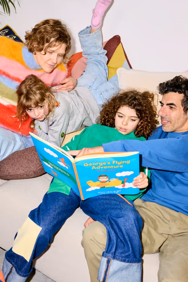 A family of four sitting on a couch reading a colorful children's book titled 'George's Flying Adventure'.