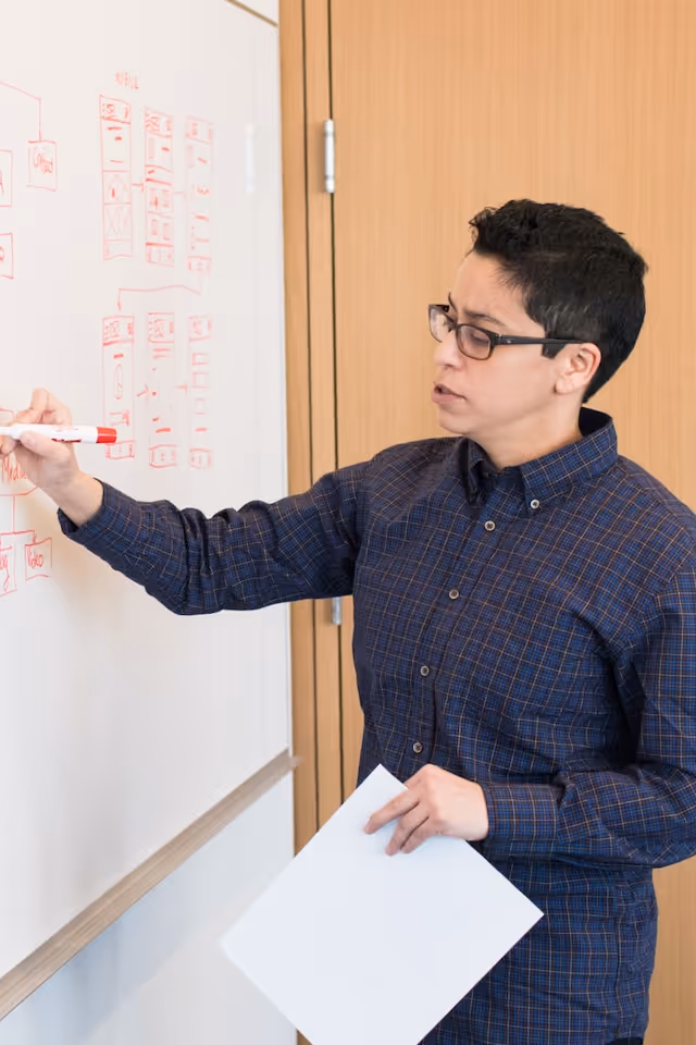 Person wearing glasses and a checkered shirt writing on a whiteboard with red marker while holding a sheet of paper.