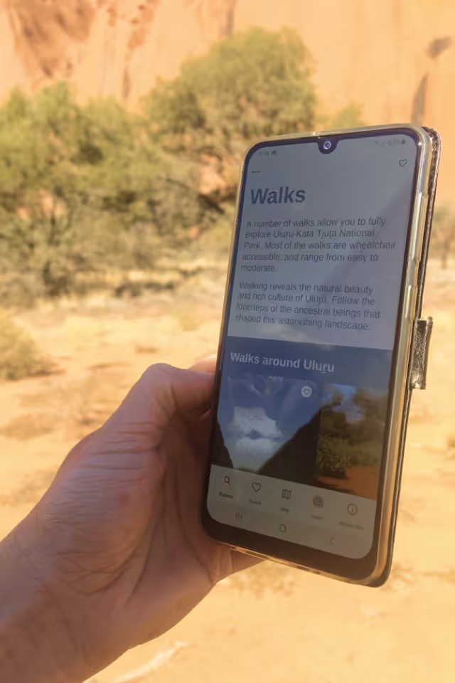Hand holding a smartphone displaying information about walks around Uluru against a backdrop of desert landscape and vegetation.