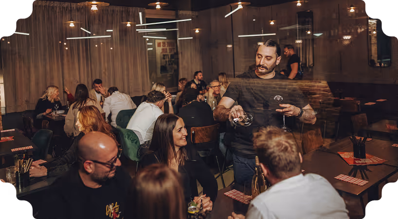 Man pouring wine into a glass at a social gathering in a warmly lit restaurant with people talking and laughing at tables.