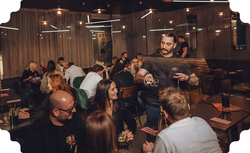 Man pouring wine into a glass at a social gathering in a warmly lit restaurant with people talking and laughing at tables.