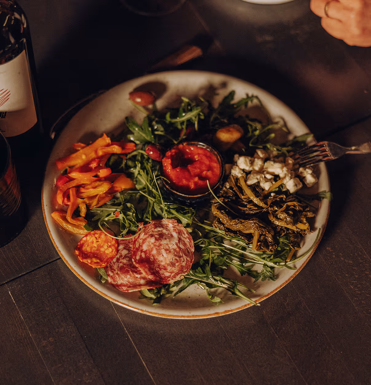 Plate with arugula, sliced cured meats, roasted peppers, cooked greens, feta cheese, and a small dish of tomato sauce.