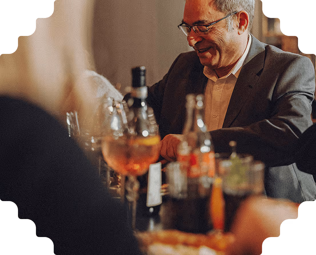 Smiling older man in glasses and suit sitting at a table with drinks in a dimly lit setting.