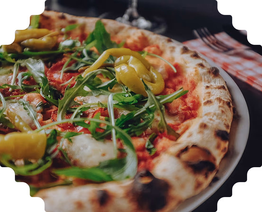 Close-up of a pizza topped with arugula and yellow chili peppers on a white plate with a fork and red checkered napkin in the background.