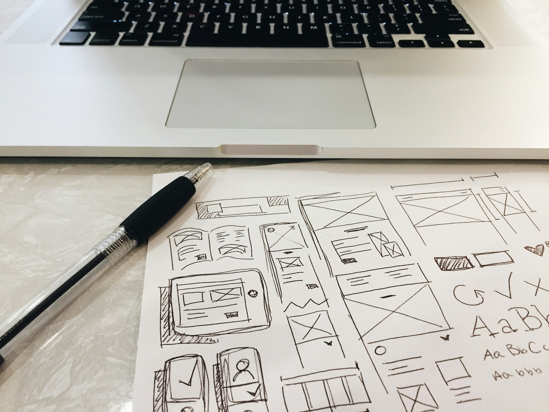 Pen and paper with hand-drawn website wireframe sketches placed in front of a laptop keyboard.