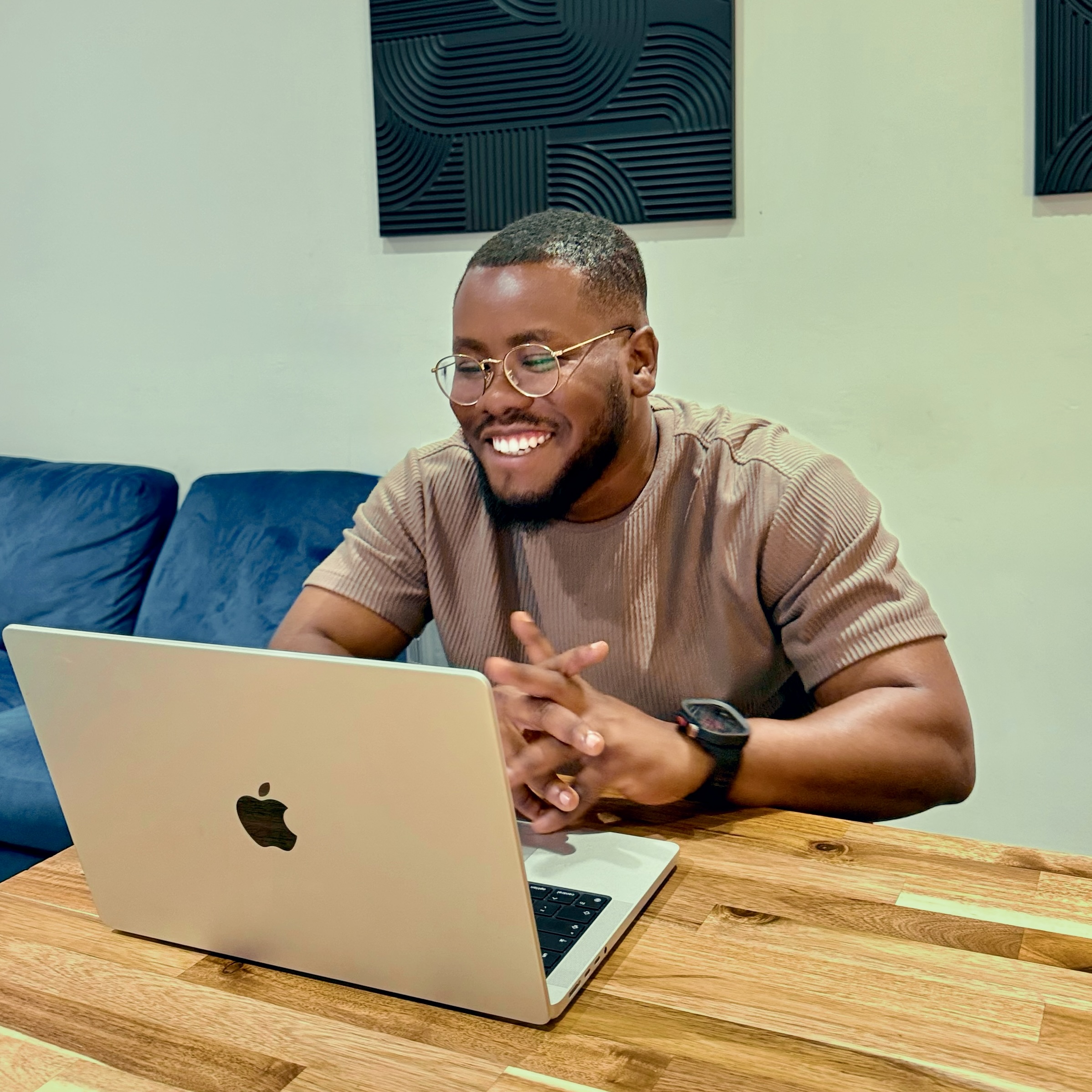 Smiling man with glasses and a beard using a laptop on a wooden table indoors.