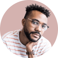 Man with short curly hair, glasses, and a striped shirt resting his chin on his hand against a light pink background.