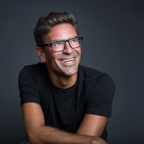 Hymyilevä mies mustassa paidassa ja silmälaseissa katsoo ylöspäin harmaata taustaa vasten.