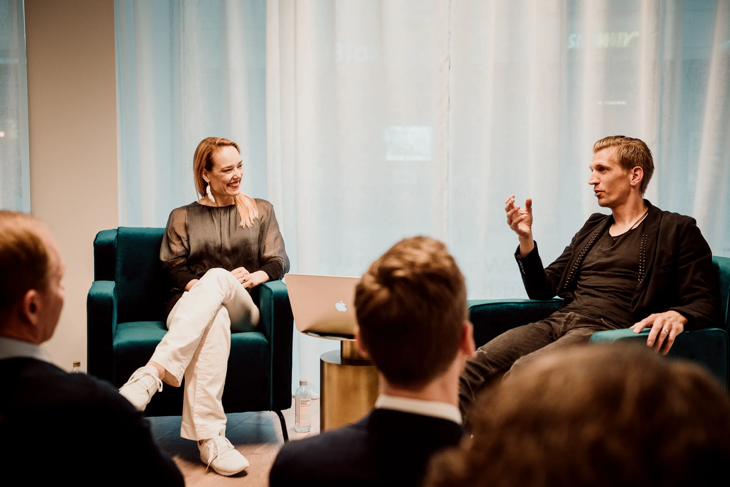 Kaksi ihmistä istuu vihreissä nojatuoleissa valkoisten verhojen edessä, keskustellen yleisön edessä. Nainen on pukeutunut harmaaseen paitaan ja valkoisiin housuihin, kun taas mies on pukeutunut mustaan takkiin ja tummiin housuihin. Huoneessa on pehmeä valaistus.