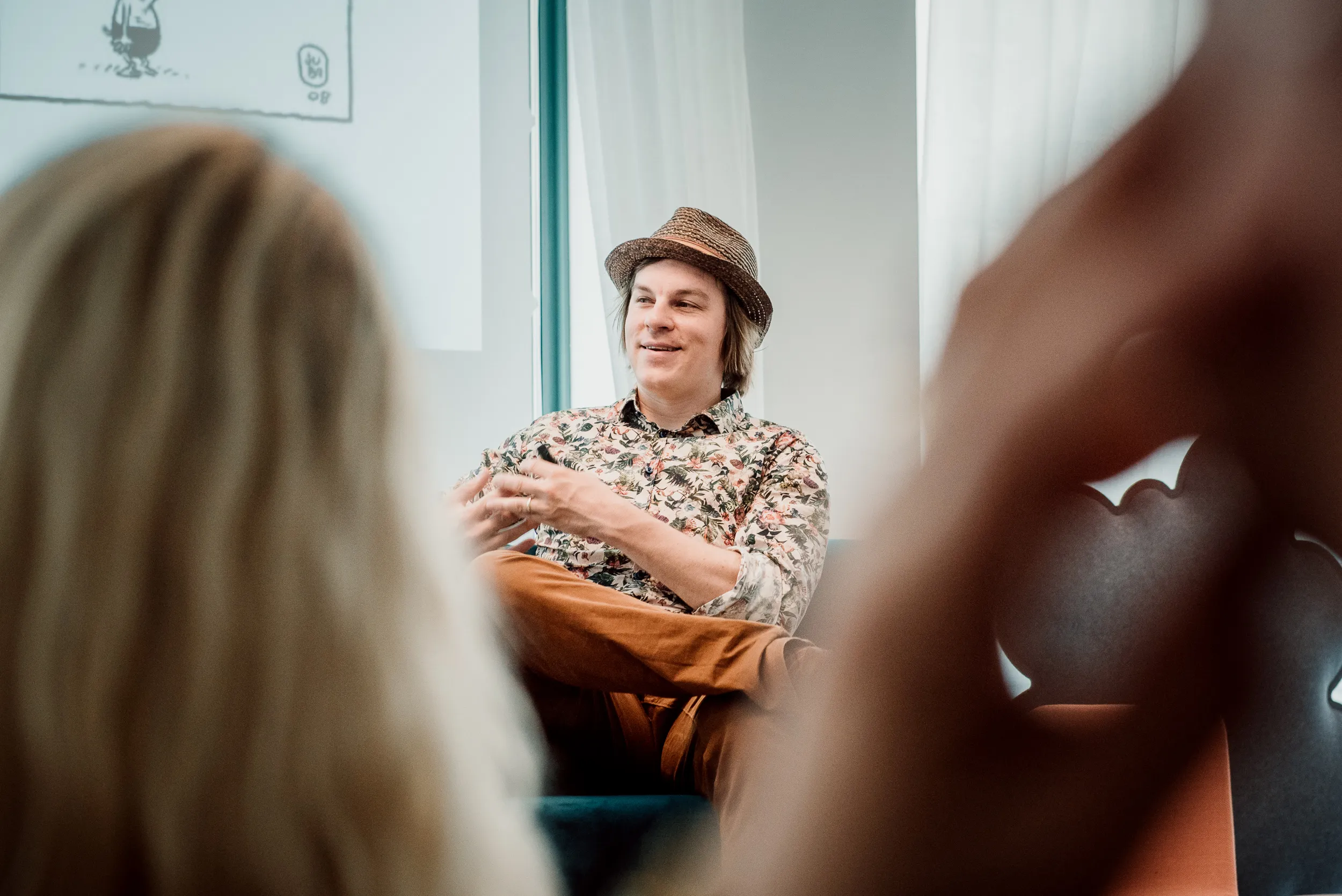 Kuvassa on hymyilevä mies olkihatussa ja kukkapaidassa, ruskeat housut jalassa, istumassa turkoosilla sohvalla. Etualalla on epätarkka henkilö ja taustalla valkoinen seinä ja ikkuna.