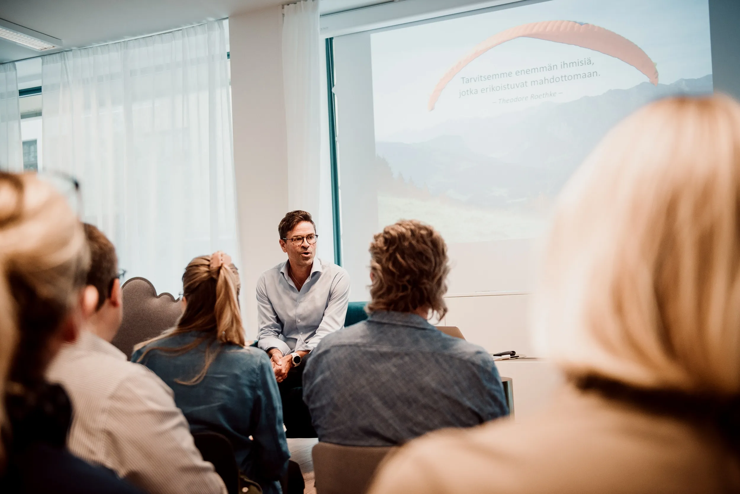 Esitelmää pitävä mies yleisön edessä, taustalla valkokangas, jossa maisemakuva ja teksti.