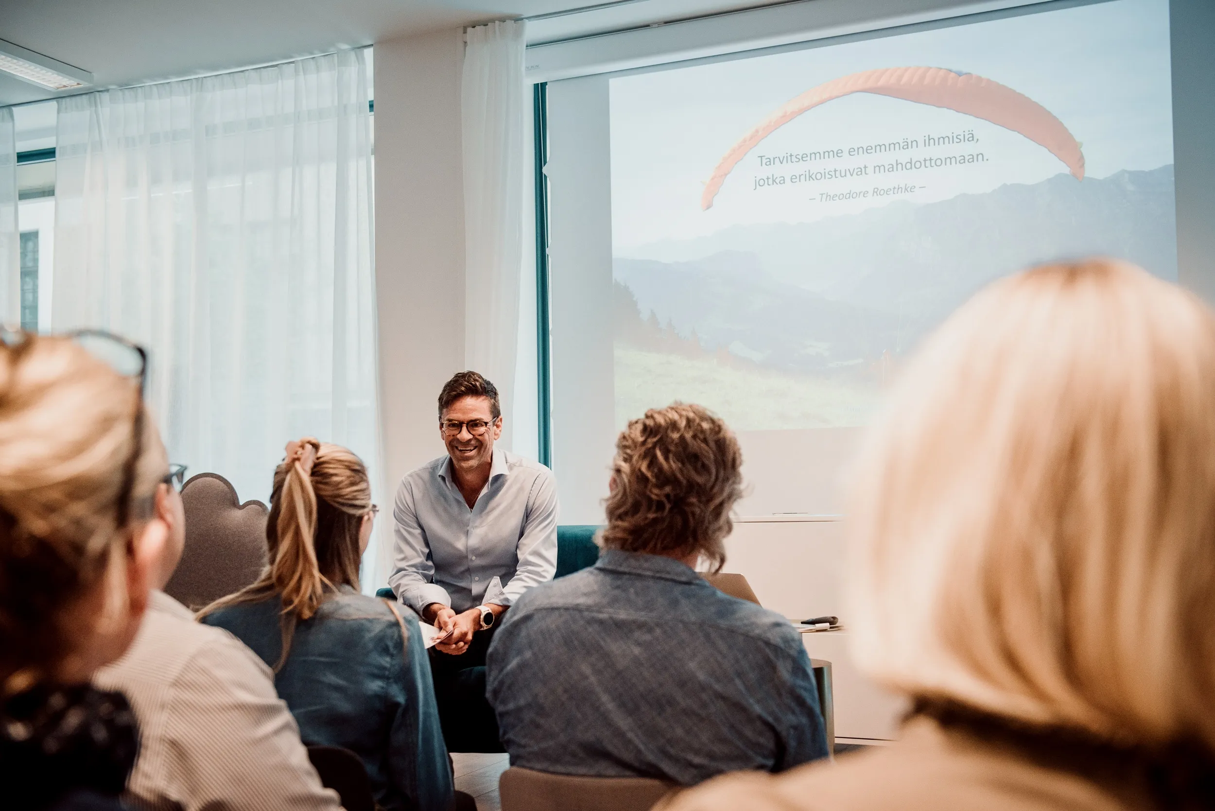 Seminaaritilanne, jossa mies esitelmöi yleisölle, taustalla valkokangas ja maisemakuva, valoisa tila.