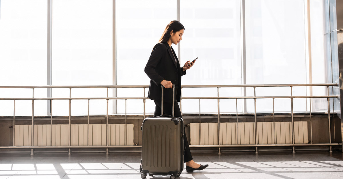 Woman in business attire walking through an airport terminal with a suitcase while checking her phone, representing travel abroad for fertility treatment.