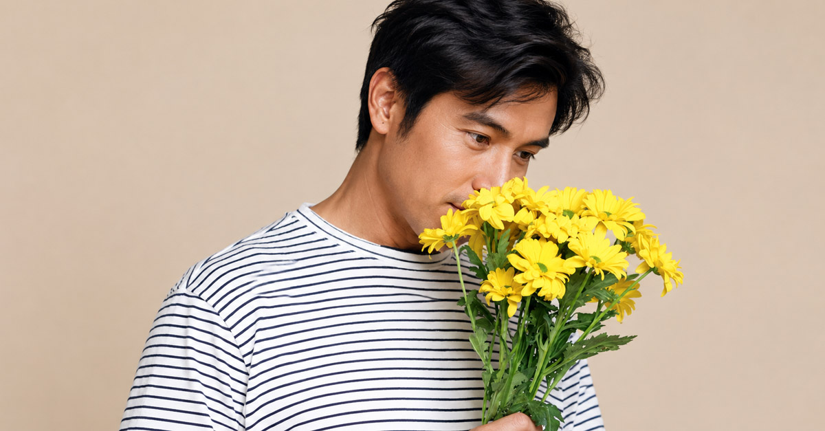 Asian man in a striped shirt holding and smelling a bouquet of yellow flowers against a neutral background, representing a male fertility health topic.