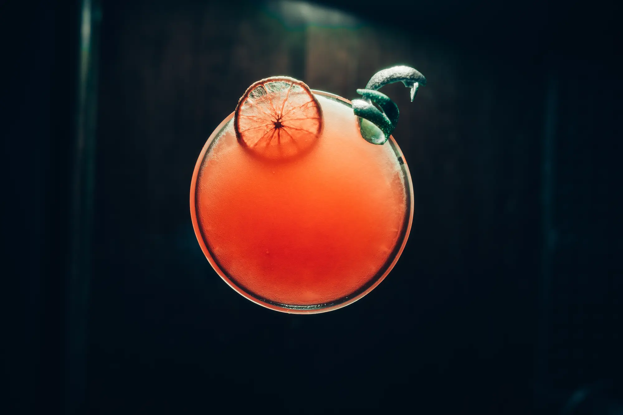 Top-down view of a red cocktail in a round glass, garnished with a dried citrus slice and mint against a dark background.