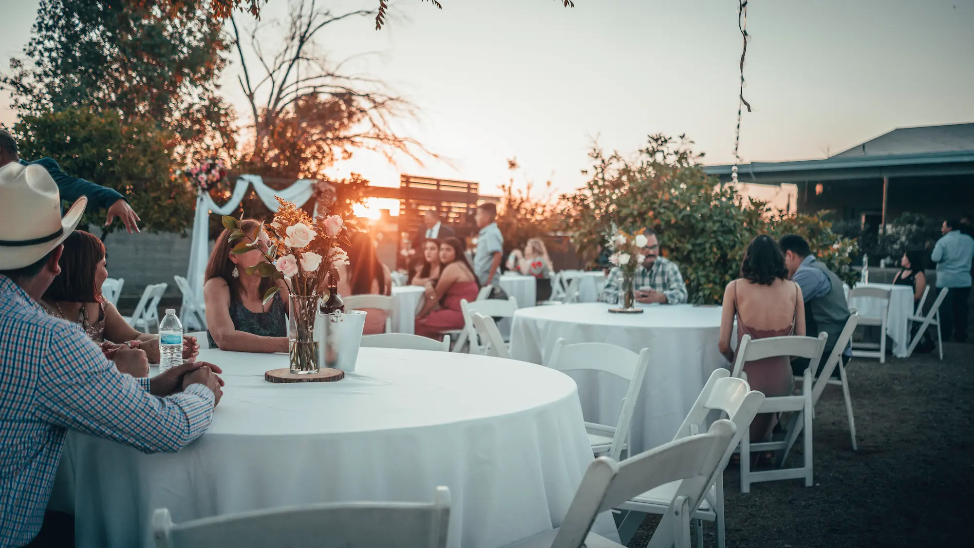 Guests seated at round tables during an outdoor event at sunset, with people socializing and decorations throughout the space.