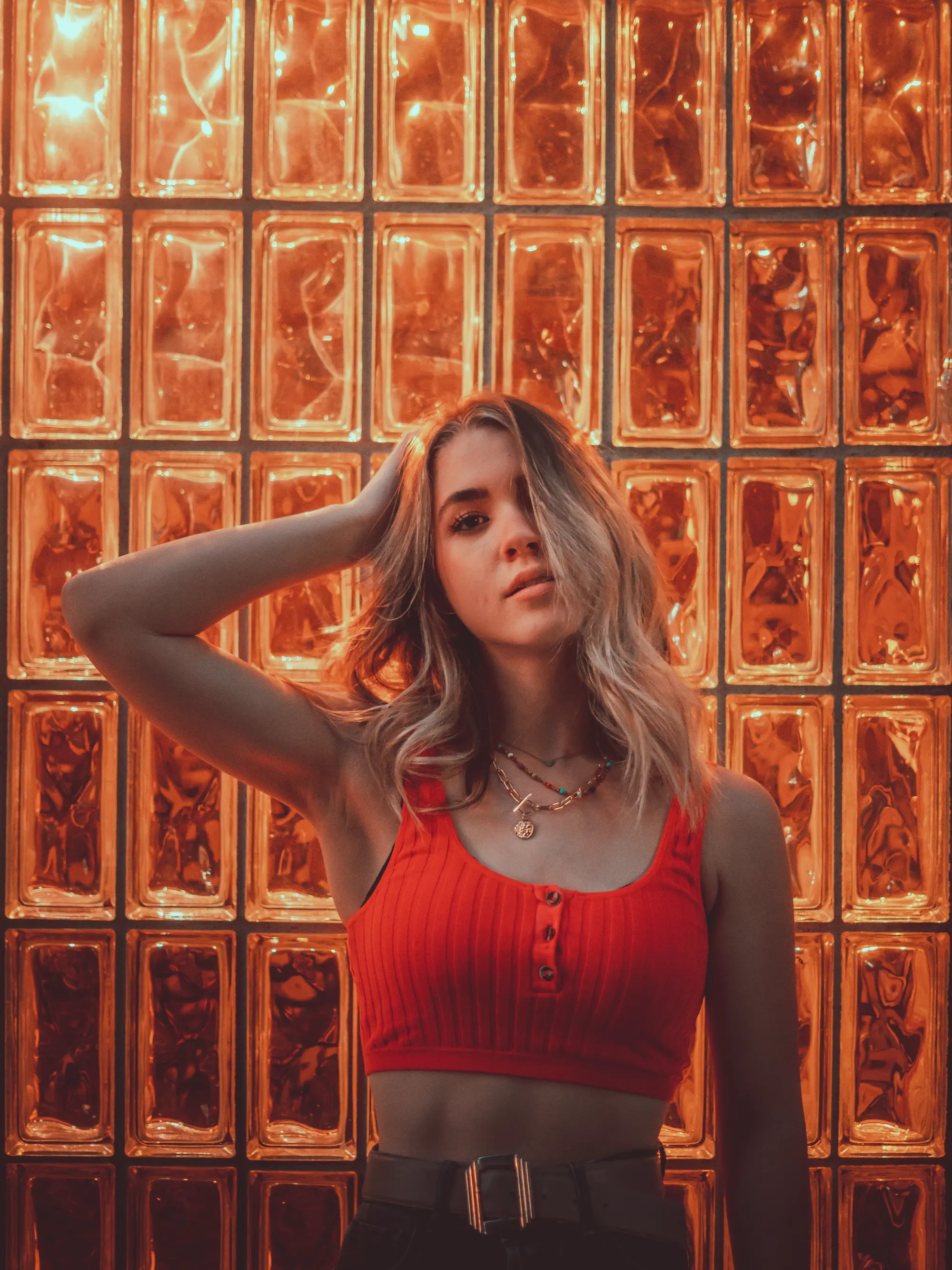 Person standing in front of an amber glass block wall, wearing a red sleeveless top and posing with one hand raised to their head.