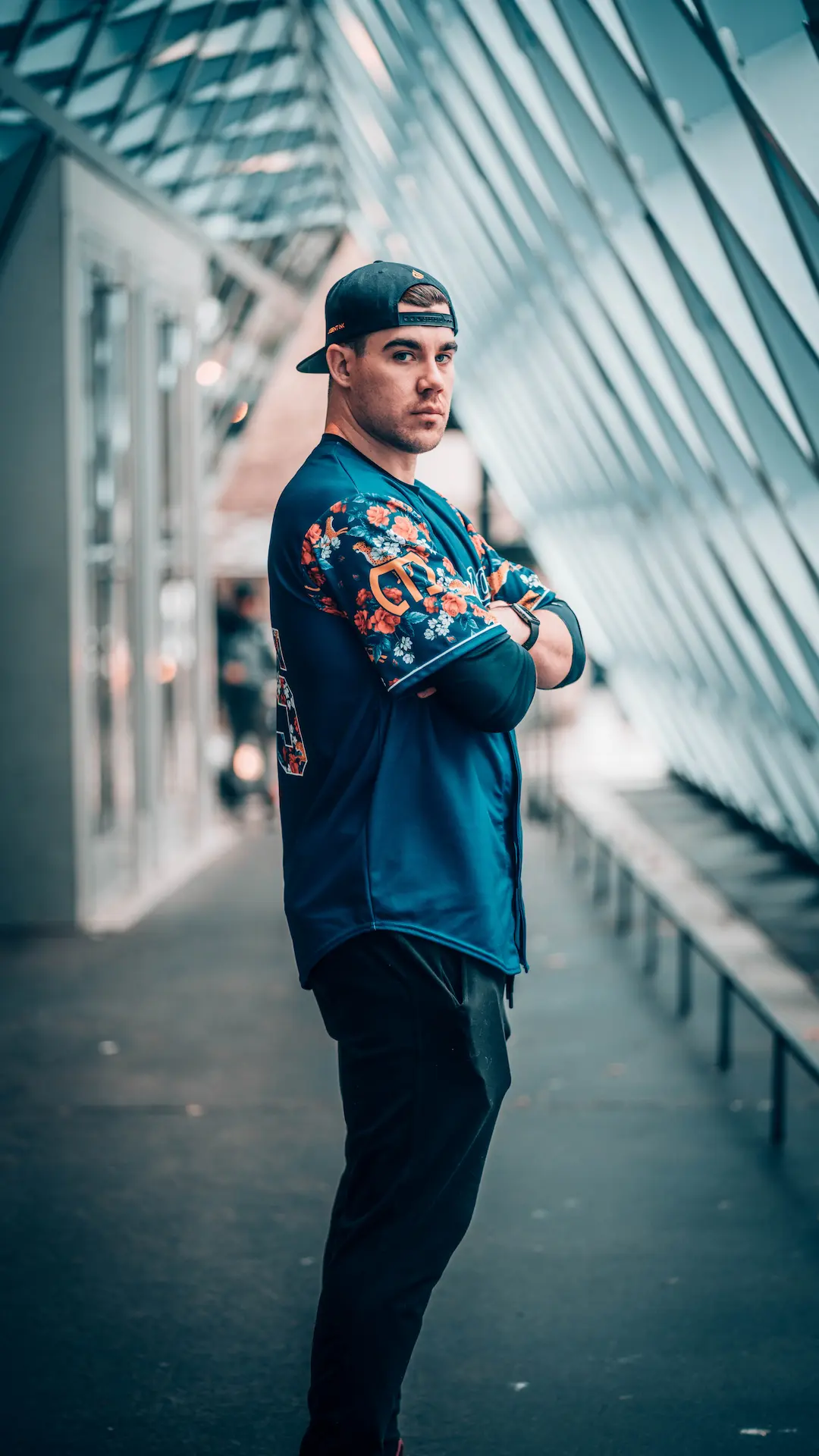 Person standing indoors with arms crossed, wearing a patterned shirt and cap in a modern, architectural hallway.