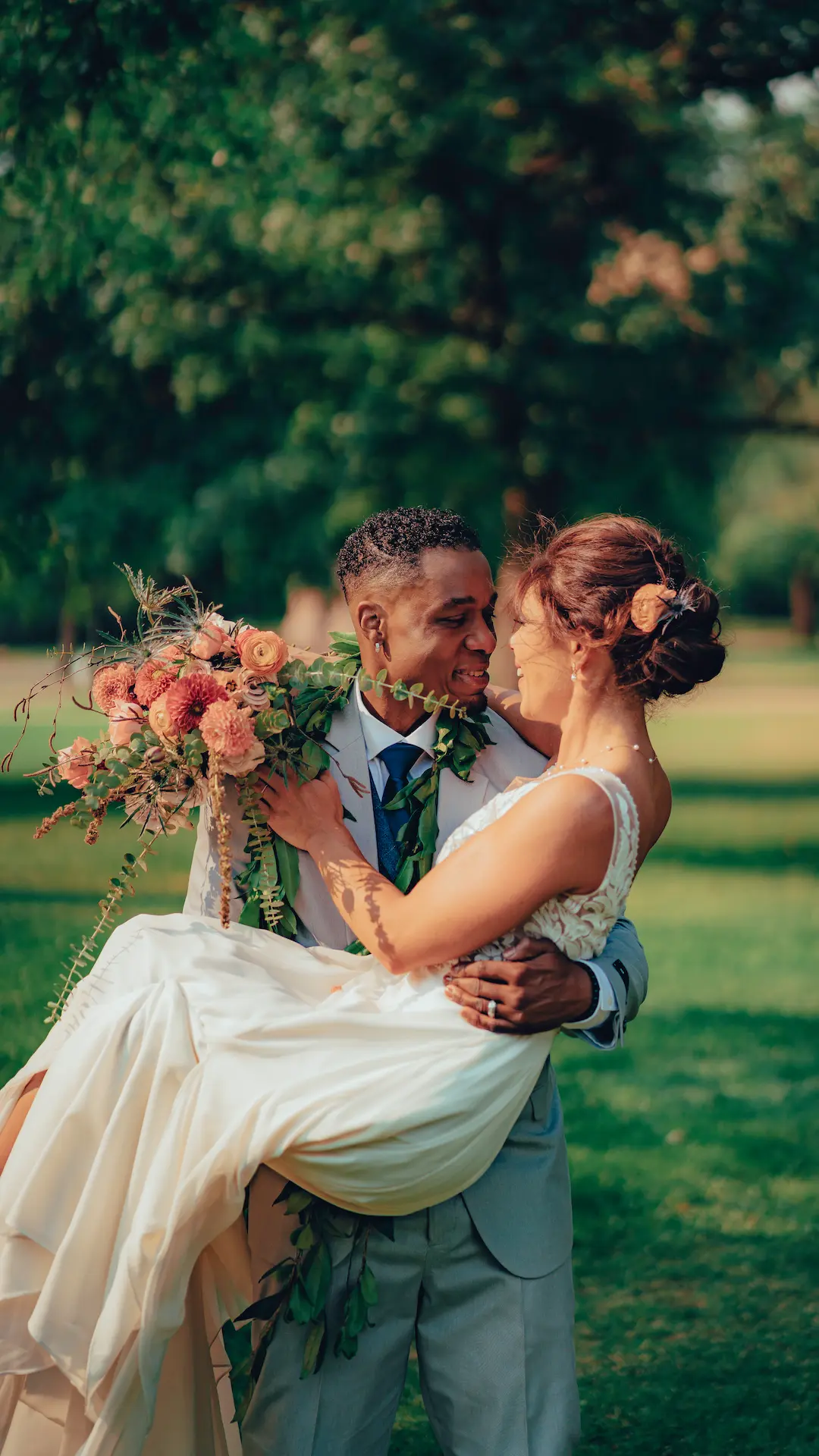 Groom lifting the bride in his arms during an outdoor wedding moment surrounded by greenery.