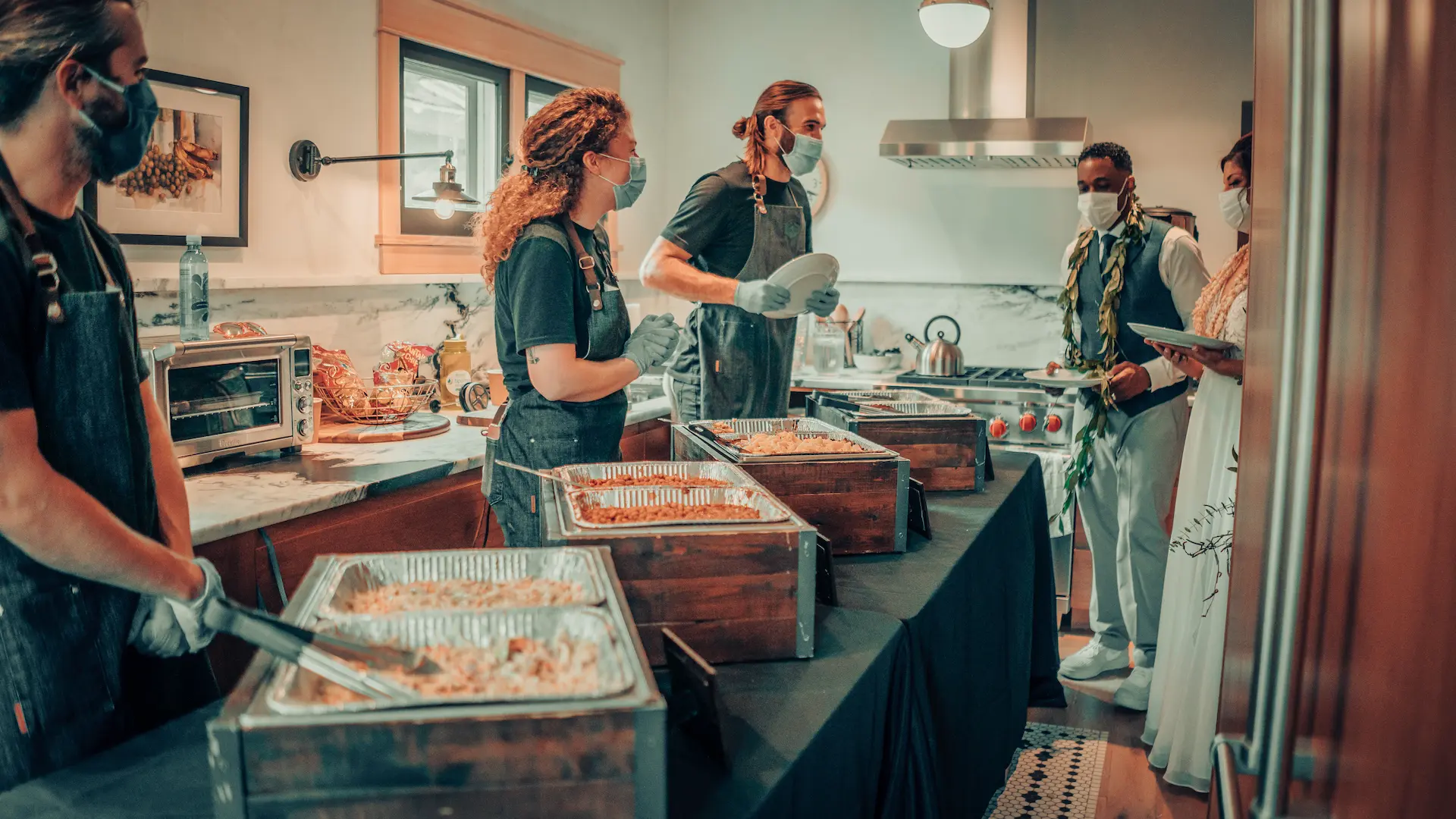 Catering staff serving food from chafing dishes at an indoor event while guests look on.