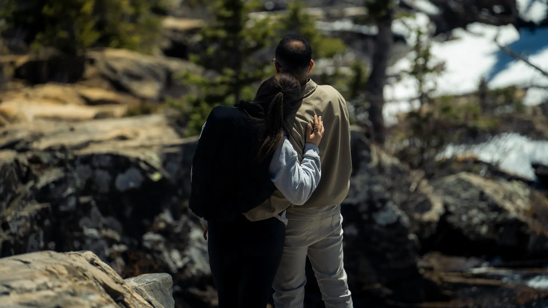 Couple standing close together on a rocky overlook, looking out at a mountain landscape.
